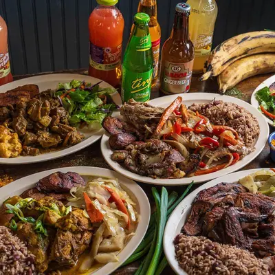 Several plates of Jamaican food laid out together with bottles of Ting and Jamaican sodas.