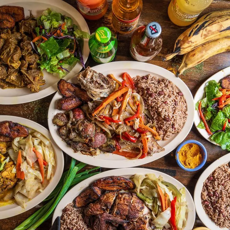 A spread of Jamaican plates, cabbage, rice and peas and bottled sodas across a wooden table.